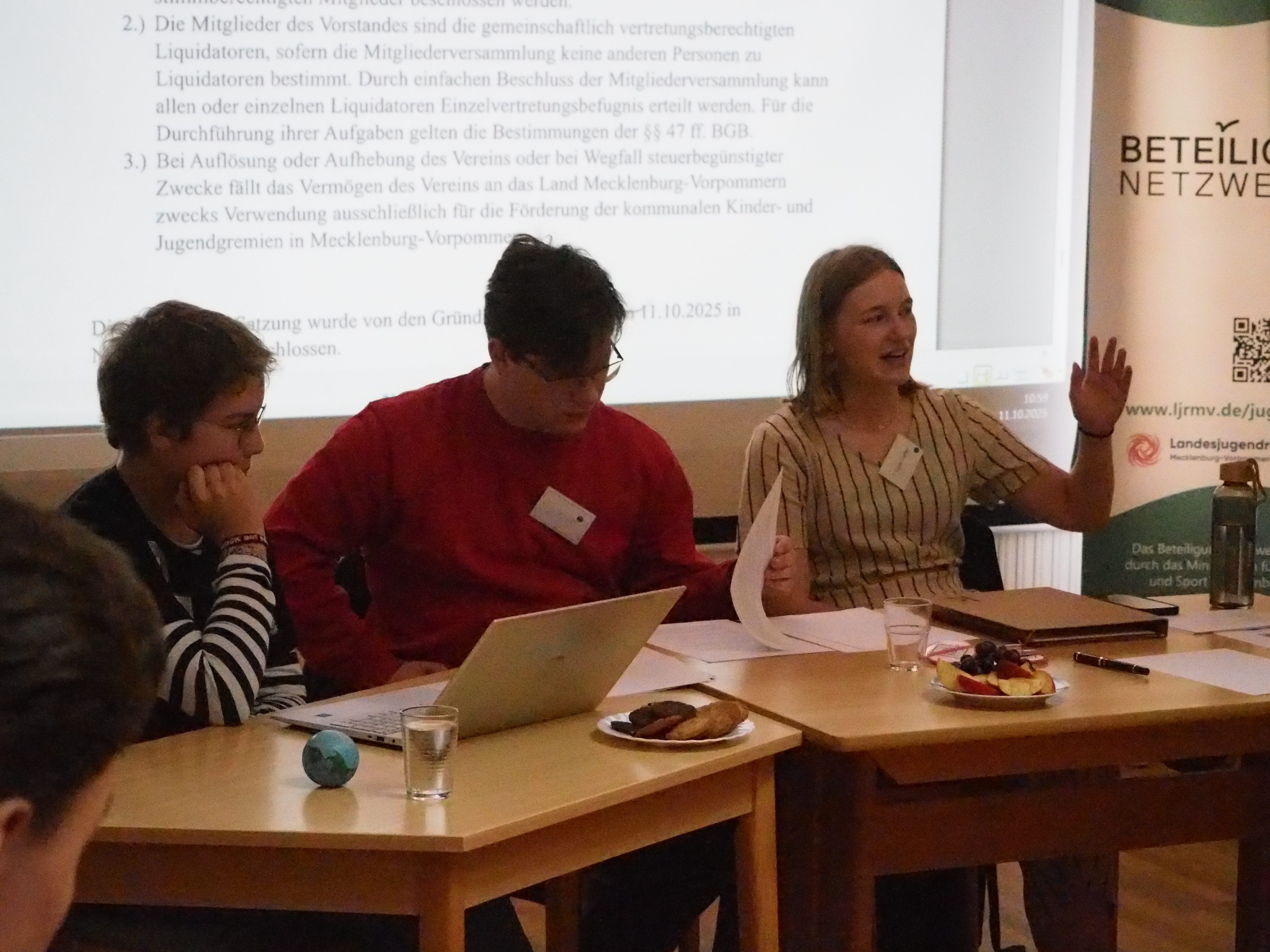 3 Jugendliche sitzen nebeneinander an zwei Tischen. Hinter ihnen ist eine Leinwand mit Text. Auf den Tischen ein Laptop, Obst, Kekse und Zettel. Die Jugendliche ganz rechts redet und hebt die Hand.